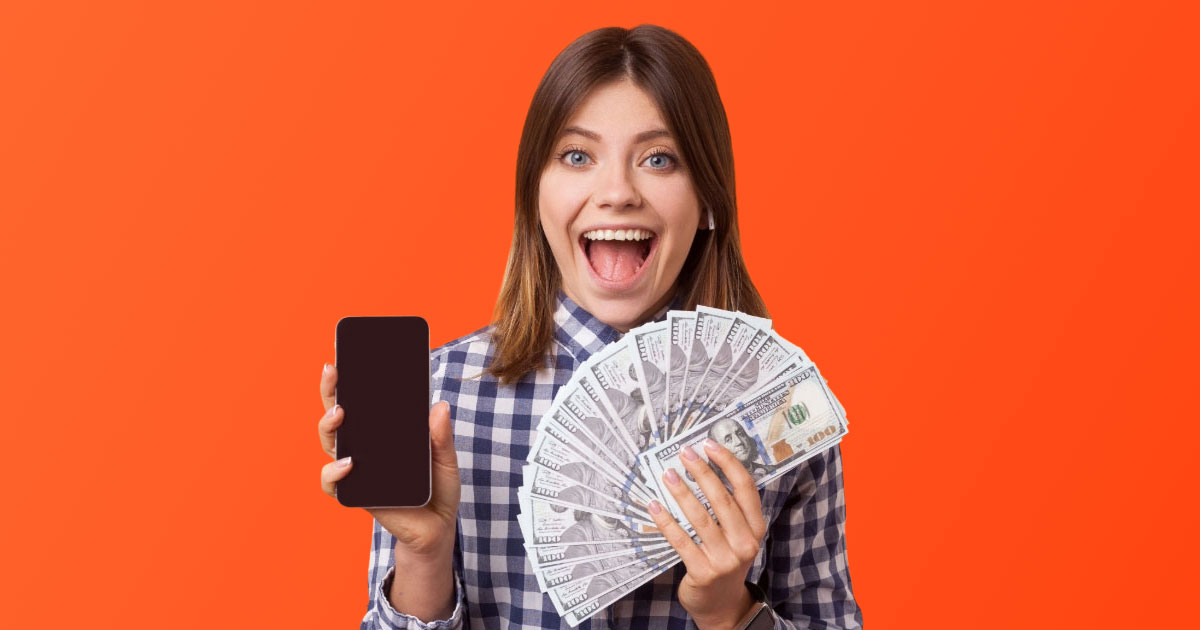 Brunette woman holding mobile phone in one hand and cash fanned out in the other on an orange background