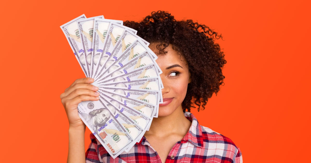Pretty woman with curly dark hair wearing red checkered shirt fanning out cash covering right side of face on orange backing